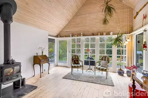 Brændeovn står varmende i stuen med lyse trægulve og et loftrum fyldt med planter. Store vinduer giver udsigt til haven. To stole og et skrivebord står ved siden af.