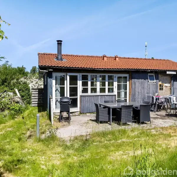 Sommerhus med rødt tegltag står stille i sollys. Foran huset er der havemøbler på en terrasse, omgivet af grøn græsplæne og træer.