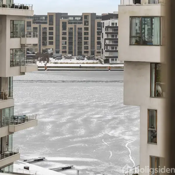 Høje, moderne lejlighedsbygninger står stille, omgivet af et vinterlandskab med en frossen sø eller havn i baggrunden. I det fjerne ses yderligere række af bygninger.