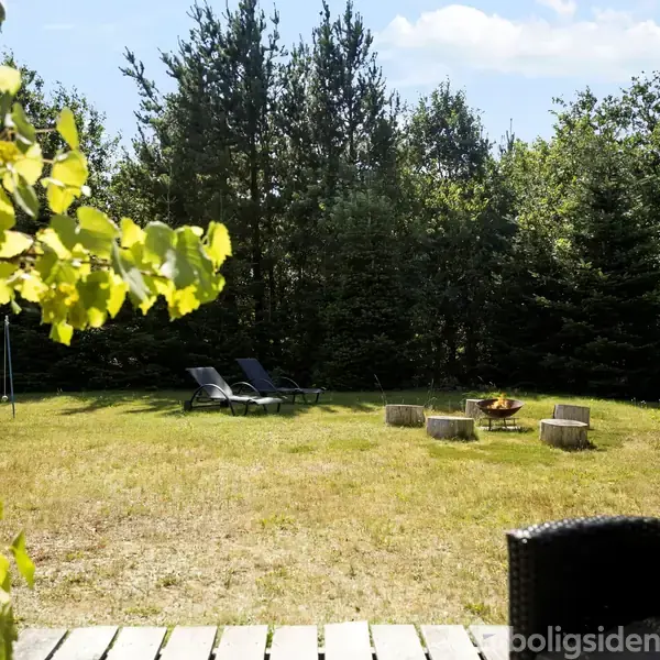Terrasse med udsigt til en grøn have, hvor to liggestole står på græsset. Træstubbe omringer et bålsted under klar himmel. Løv fra en plante hænger over terrassen.