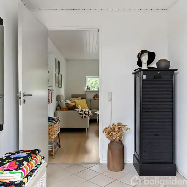 En hvid dør åbner ind til en stue med en beige sofa og farverige puder. En høj, sort kommode står ved døren med en skulptur og stråhat ovenpå.