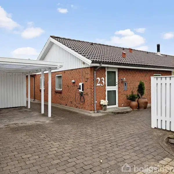 En etplans murstensvilla med nummer 23 på væggen har en carport til venstre og en flisesti foran indgangen. Omgivet af grøn hæk og blå himmel.