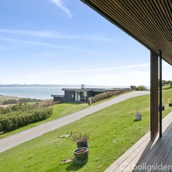 Terrasse med udsigt over en græsplæne og en kystlinje. Flere potteplanter står på terrassen. I baggrunden ses et moderne hus og en vej, der fører til havet.