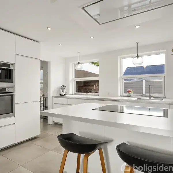 Et moderne køkken med en stor, hvid køkkenø og indbyggede hvidevarer. To sorte barstole står ved øen. Store vinduer giver naturligt lys, og en blomstervase pynter bordet.