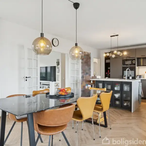 Spisebord med stole står i et lyst køkken-alrum med hængende lamper. Køkkenet har moderne skabe og en stor ø, omgivet af træmøbler og hvide vægge med en enkelt pendel.