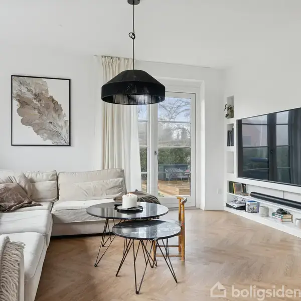 En lys stue med en hvid hjørnesofa foran et stort vindue. To små, runde borde står foran sofaen. En stor fladskærm er monteret på væggen til højre.