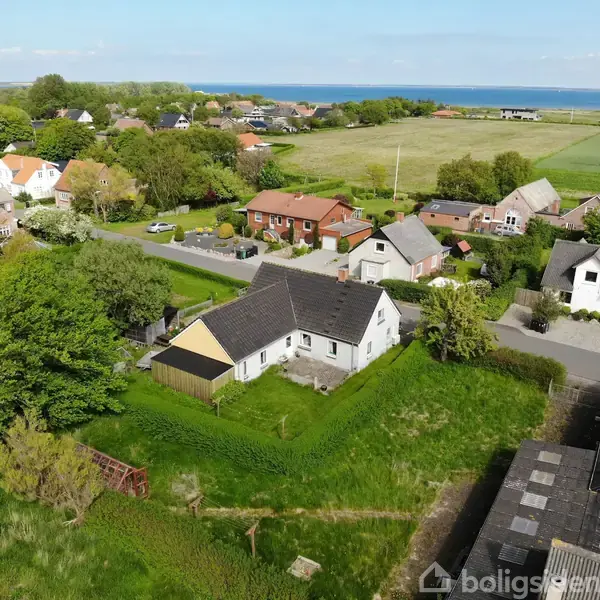 Hus med sort tag og hvid facade ligger i en frodig have. Det er omgivet af andre huse og grønne områder med havudsigt i baggrunden.
