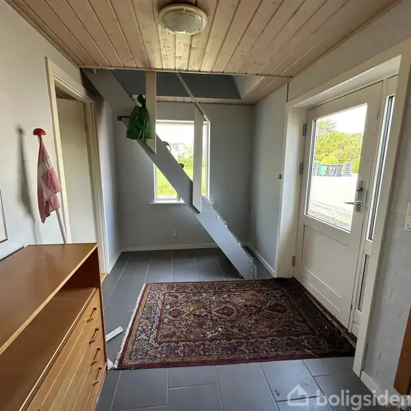 Gulvtæppe ligger i en entré med gråt flisegulv. Trappe opad til venstre. Kommode står nær væg med farverigt kunstplakat. Dør og vindue til højre.