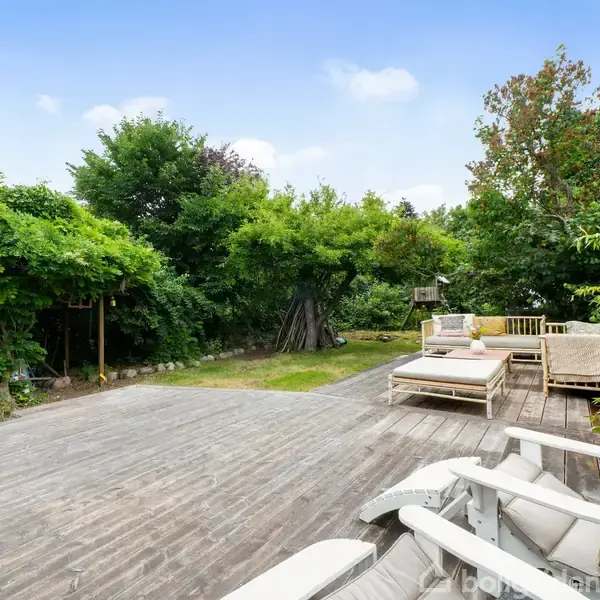 Terrasse med træmøbler og en sofa står stille, omgivet af frodig have med grønne buske og træer. En græsplæne strækker sig ved siden af terrassen.