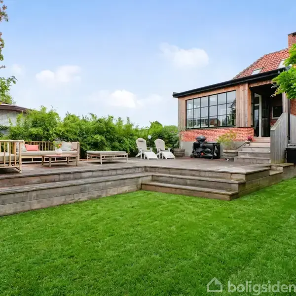 Hus med rød mursten og terrasse i træ har liggestole og sofa. Omkring er der velplejede plæner og buske, mens solen skinner fra en let skyet himmel.