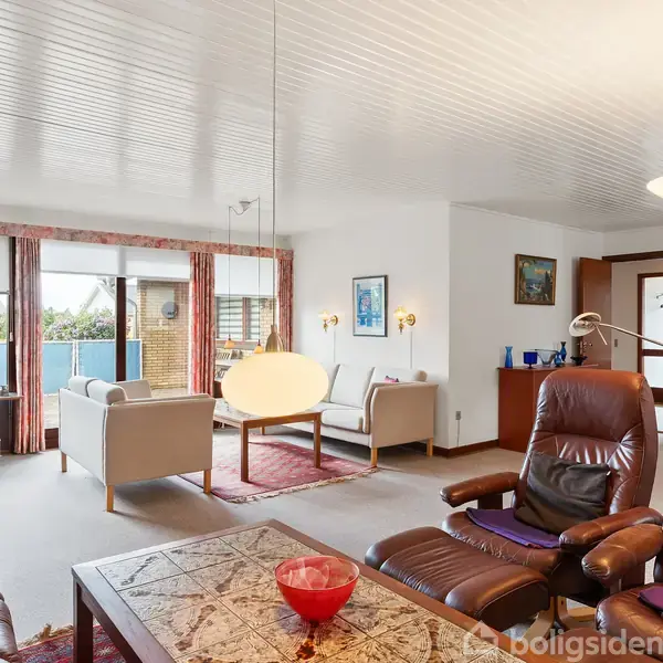 Stue med brune lænestole og et sofabord. Store vinduer med gardiner lukker lys ind. Rummets dekoration inkluderer billeder og lamper. Elegante møbler skaber en indbydende atmosfære.