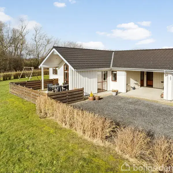 Hus med sort tag og hvid facade står stille ved grusvej, omgivet af have og græsplæne med buske. Danmarksk landskab omkranser ejendommen med træer og åbne marker.