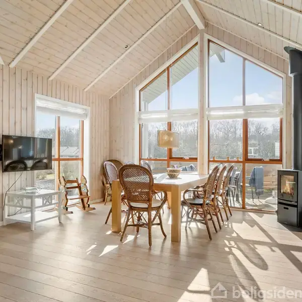 Et lyst træbeklædt rum med store vinduer. Bord med stole i midten, pejs til højre. Moderne interiør og udsigt til natur udenfor.