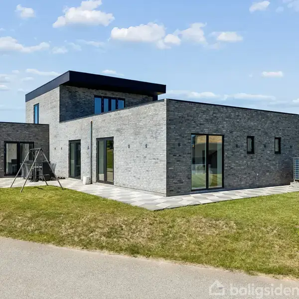 Et moderne, kubisk hus med fladt tag står på en græsplæne. Store vinduer præger facaden, og en lille terasse omgiver huset. Himlen er klar med få hvide skyer.
