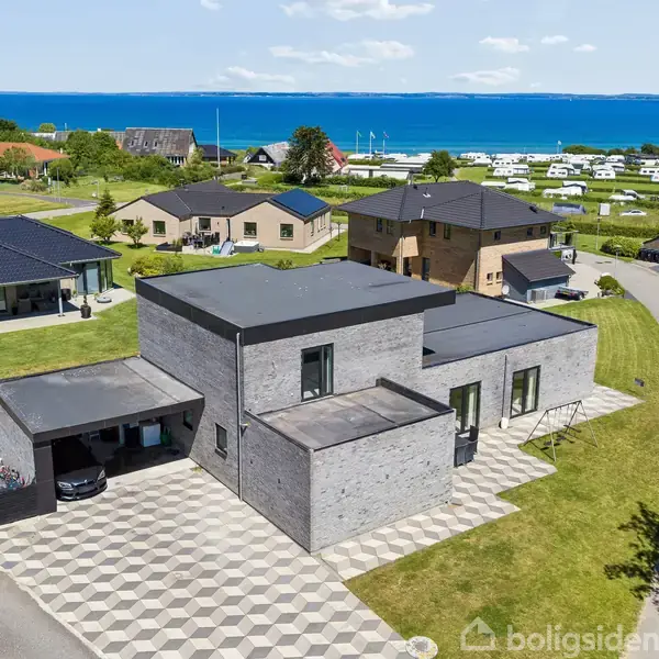 Moderne grå murstenshus med fladt tag står på en flisebelagt indkørsel. Baggrunden viser flere huse, en campingplads og et blåt hav under en klar himmel.