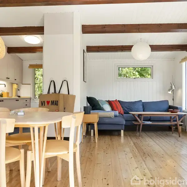 Stue med blå sofa og farverige puder står ved vindue. Spisebord med stole foran. Køkken i baggrunden. Træloft med synlige bjælker. Lampe hænger over bordet. Papirpose med tekst "VI ♥ MAR".