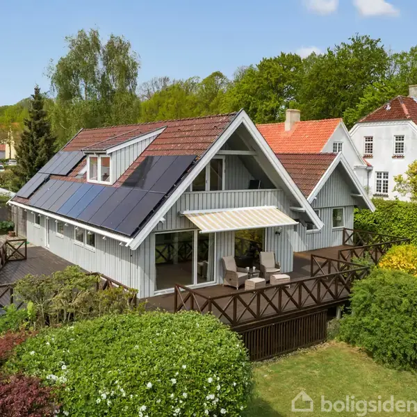 Et hvidt hus med træbeklædning har solpaneler på taget. Det står i en frodig have med terrasser omgivet af buske og træer. Andre huse er synlige i baggrunden.