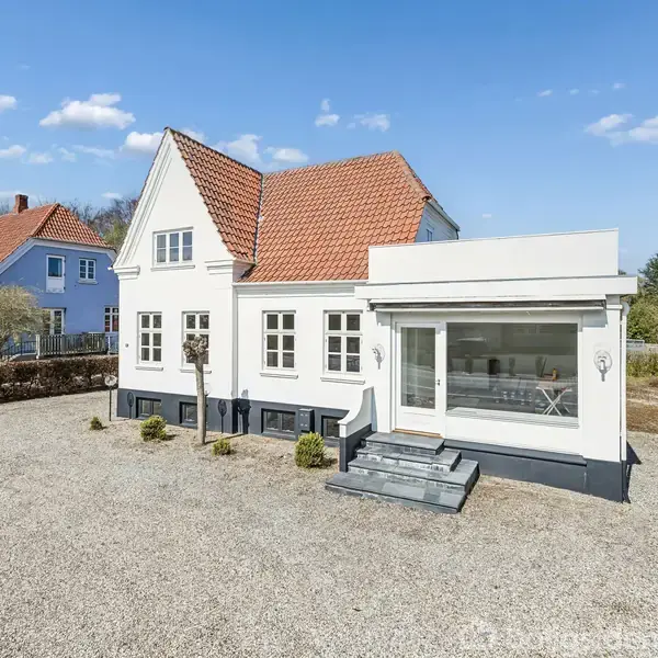 Hvidt hus med rødt tegltag, stående stille på en grusbelagt gårdsplads, omgivet af træer og buske, med en blå himmel i baggrunden.