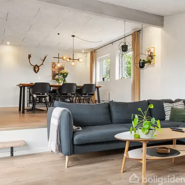 En moderne stue med en grå hjørnesofa og et rundbord med planter foran. I baggrunden ses en spisestue med et stort bord og stole, omgivet af lyse vægge og vinduer.