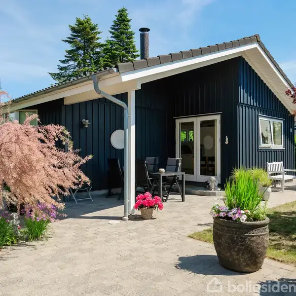 Sort træhus med terrassemøbler, stående på en skærvet gårdsplads. Omkring er der blomsterkrukker og spredte farverige buske. Himlen er blå og omgivelserne er frodigt beplantet med træer.