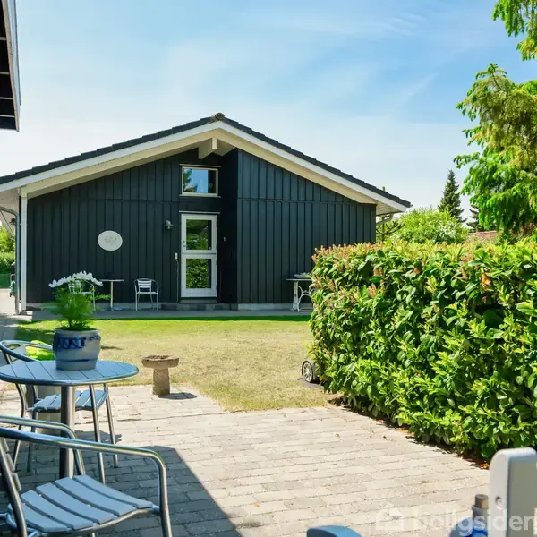 Et sort hus med store vinduer står i en have. To stole og et bord ses nær buske ved stenterrasse. Græsplænen og træer omkranser området.