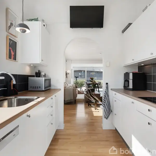 Et moderne køkken med hvide skabe, sort kvadratisk flisevæg og træbordplader. I baggrunden ses en lys stue med vinduer. Sollys strømmer ind fra venstre.