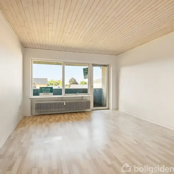 Stue med lyst trægulv har store vinduer og dør mod balkon, hvor der ses udendørs træer. På vinduet står teksten: "Til salg" og "EDC Ejendoms Centret".