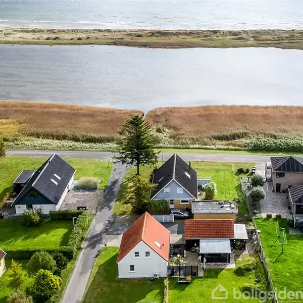 Huse ligger tæt på en vej langs en kyst med et vandløb og strand i baggrunden. Grønt græs og træer omkranser området, under en klar himmel.