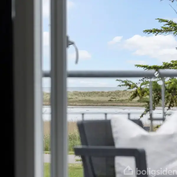 Stol med en hvid pude står på en terrasse, der vender ud mod en sø eller flod med grønne træer og en åben, blå himmel i baggrunden.