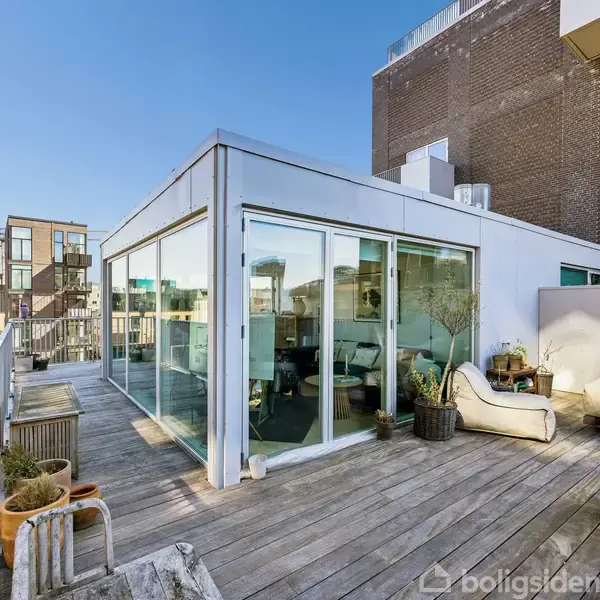 En moderne taglejlighed med store glaspartier. På terrassen er der loungemøbler og potteplanter. Omgivet af byens skyline med bygninger i baggrunden under en klar, blå himmel.
