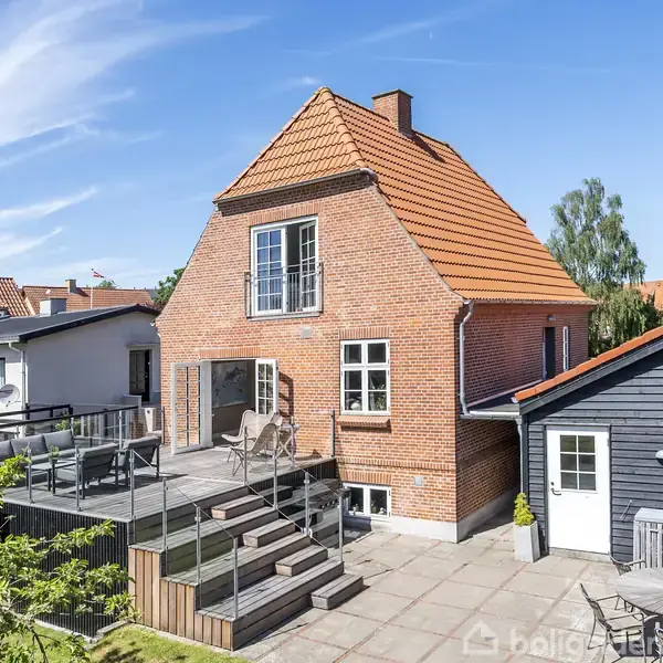 Rødt murstenshus med rødt tegltag omgivet af en solrig terrasse med trapper. Haven har græs og buske. Der er tilstødende grå træbygning. Klar blå himmel i baggrunden.