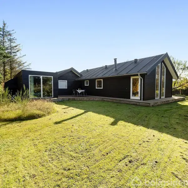 Sort træhus med store vinduer står i solbeskinnet have omkranset af træer og græs.