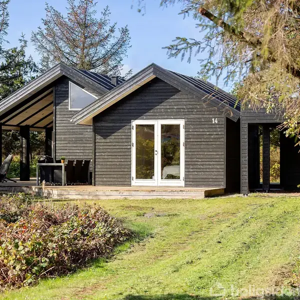 Et sort træhus med store vinduer står i en grøn have omgivet af træer og buske. Nummeret "14" hænger ved døren, og en terrasse er synlig foran huset.