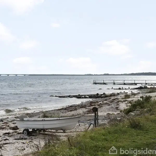 Båd hviler på sandet ved kystlinjen, omgivet af hav, tang og en fjern bro. En grussti og en mole ses også langs stranden.
