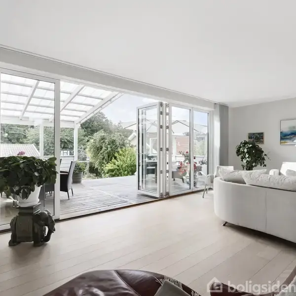 En stue med hvid sofa står foran store, åbne glasdøre, der fører til en overdækket terrasse med spisebord. Rummets lyst og moderne indretning er indrammet af planter.