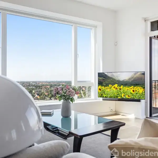 Stue med en hvid lænestol og sofabord. Et fjernsyn viser et bjerglandskab med solsikker. Store vinduer og altandør åbner til en udsigt over byen. Blomsterdekoration på bordet.