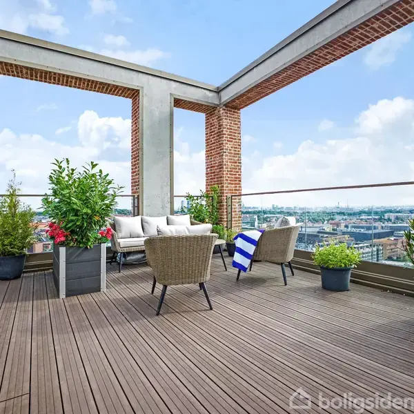 En tagterrasse med sofa og stole står blandt planter, der omgiver et åbent område med udsigt over byen. Himmel med skyer.