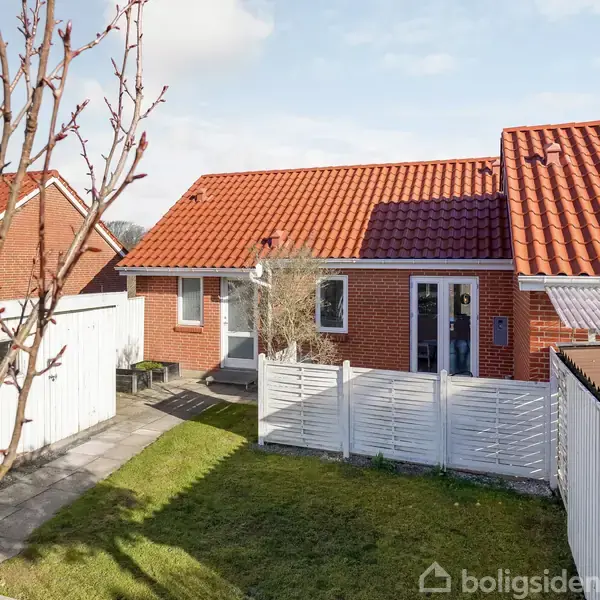 Rødt murstenshus med rødt tegltag, omgivet af en lille have og hvidt stakit. Husets dør og vinduer vender mod en stenbelagt gangsti. Ingen tekst findes på billedet.