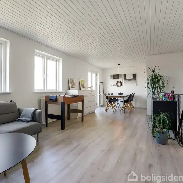 En stue med grå sofa til venstre, spillebord i midten, og spisebord med sorte stole i baggrunden. Lyse trægulve og hvidt loft. Store vinduer giver lysindfald. Plakater på væggene.