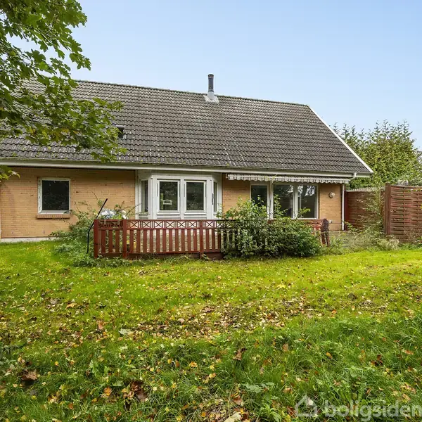 Et murstenshus med en brun facade og skråt tag ligger i en have med græs og træer. En rødlig træterrasse er foran huset, omgivet af buskads.