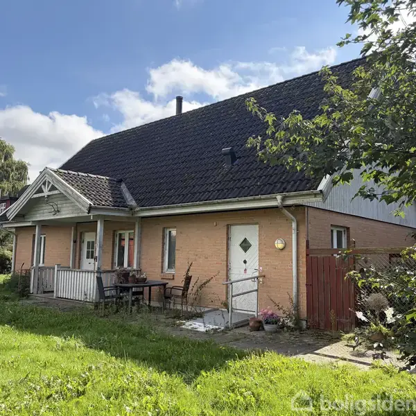 Et murstenshus med sort tag står stille i en grøn have. To indgangsdøre og en lille veranda er synlige, omgivet af træer og blå himmel.