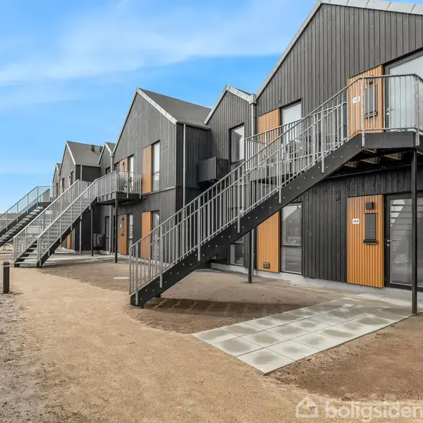 Flere moderne, sorte rækkehuse med træpaneler har udendørs trapper, der fører til den øverste etage. Omkring dem er der en sandet grund og en flisebelagt sti i et åbent område.