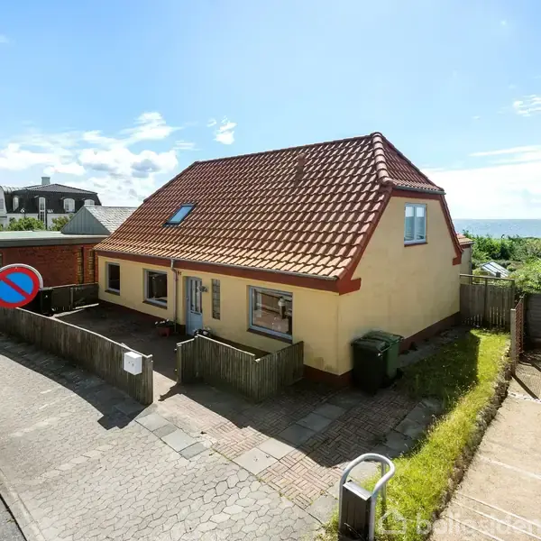 Et gult hus med rødt tag står stille ved en rolig vej. Omkring huset er der små haver og en udsigt til havet. Et skilt viser parkering er forbudt.