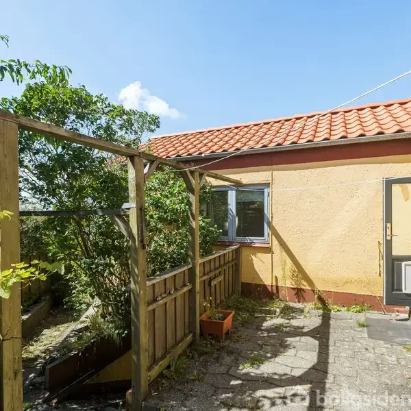 Træpergola står i en gårdhave ved et hus med gult puds og rødt tegltag. Et lille vindue og en dør er på husmuren. Sollys skaber skygger på flisebelægningen.