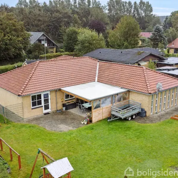 En gulmurstensvilla med rødt tag står omgivet af en grøn have. En bil er parkeret på indkørslen. Der er et gyngestativ i haven og et halvtag ved huset.