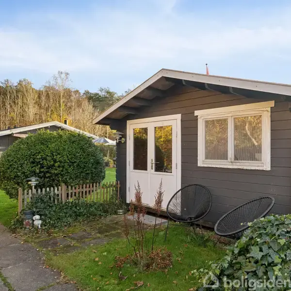 Lille træhus står stille i en have med græsplæne og buske omkring. En flisebelagt sti fører til huset. I baggrunden ses et større hus og skovbevoksning.