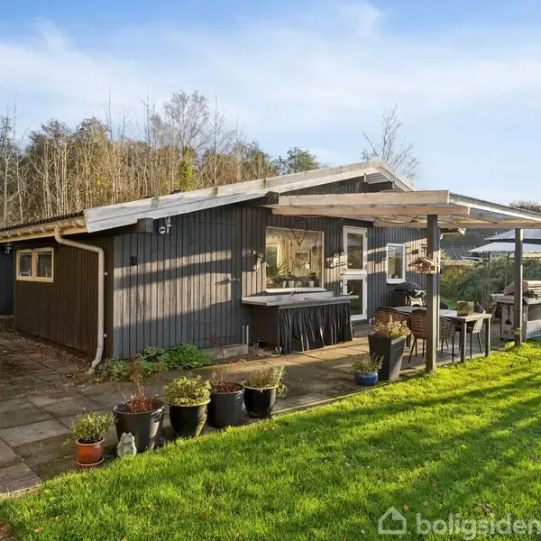 Sort træhus med terrasse, omgivet af grøn græsplæne og skov i baggrunden. Flere blomsterkrukker står på fliser langs huset, og et enkelt bord med stole er under den overdækkede terrasse.
