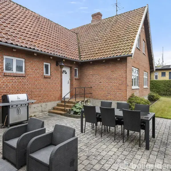 Rødstenshus med terrasse, hvor sorte havemøbler og en grill er placeret. Omkring huset er der en grøn græsplæne og en hæk, der skaber en hyggelig udendørs atmosfære.
