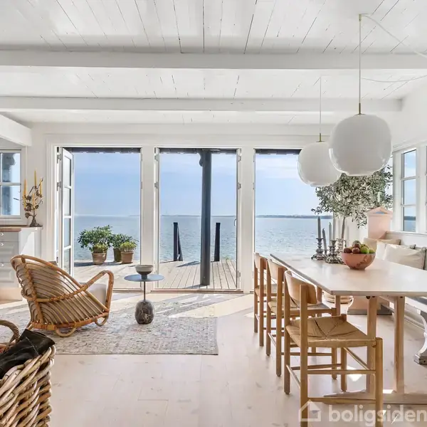 En lys stue med spisebord er udstyret med træstole og store pendellamper. Udenfor er der udsigt til havet gennem store glasdøre. Hyggelig stemning med brændeovn og kurvemøbler.