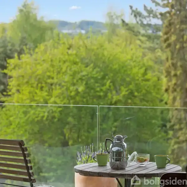 En rund træbord med en kaffekande og to kopper står på en altan. To stole vender mod en udsigt af grønne træer under en blå himmel.